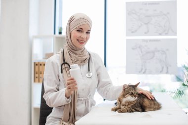 Laboratuvar önlüğü giymiş tesettürlü ve steteskoplu gülümseyen kadının portresi. Sevimli kediyi okşarken elinde bir kutu hap tutuyordu. Neşeli Arap veteriner doktor kedi hayvanlar üzerindeki pire istilasını önlüyor..