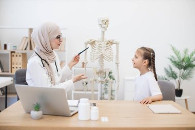 Gündelik giyinen Kafkas ergen kız klinikteki doktorların iş yerinde insan eğitim modelinin göğüs kemiğine dokunuyor. Türbanlı Müslüman doktor göğüs kemiğinin vücut fonksiyonlarındaki önemini açıklıyor.