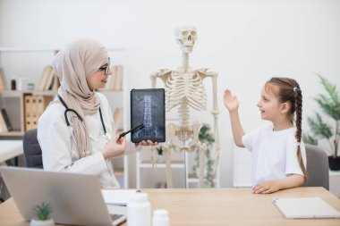 Zeki küçük çocuk doktorların işyerindeki iskeleti işaret ederken Arap tesettür uzmanı tabletteki MRI görüntüsünü inceliyor. Kafkasyalı liseli kızlara kemik anormallikleri bulmalarını öğreten bir tıp uzmanı..