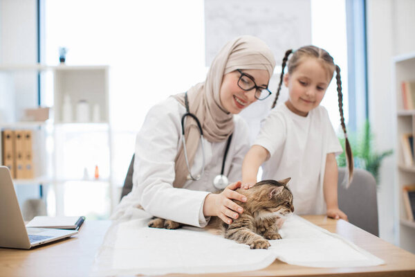 Cheerful muslim lady in headscarf and caucasian preteen girl stroking tabby cat on underpad in doctors office. Excited vet and owner helping pet to relieve stress after exam by praising and patting.