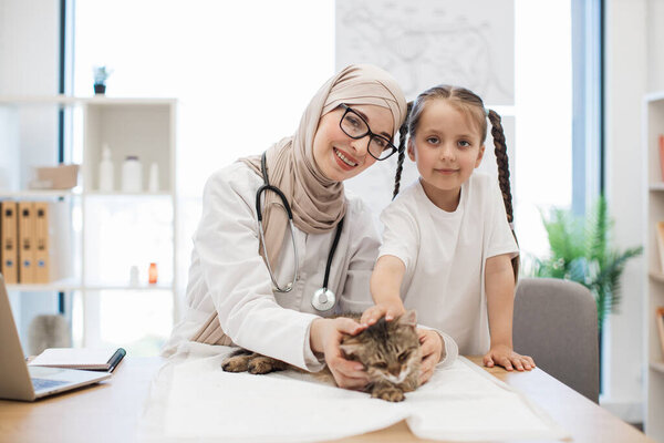 Portrait of muslim woman in hijab and smiling caucasian child posing with adult cat in doctors workplace. Peaceful furry friend relaxing on wooden desk near laptop in modern veterinary practice.