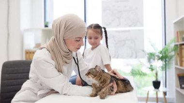 Arap uyruklu deneyimli bir kadın veteriner, sahibiyle birlikte tüylü kediyi inceliyor. Doktor steteskopla ciğerlerini dinlerken küçük kız tıbbi kanepede oturuyor..