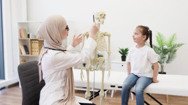 ヒジャーブの女性は骨格モデルの肋骨を示し 病院のコンサルティングルームで慎重な乳児を産みました アラビアのヘルスケア 傷害後の痛みの下で人体の部分を視覚化する専門家 — ストック動画
