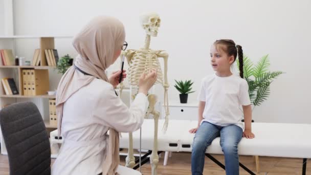 小児科医の医師室で骨格モデルの肋骨を指している白人少女の間の視界を閉じます ヒジャブにおけるアラビアの一般開業医 人間の身体の内部の働きについてコンサルティング — ストック動画