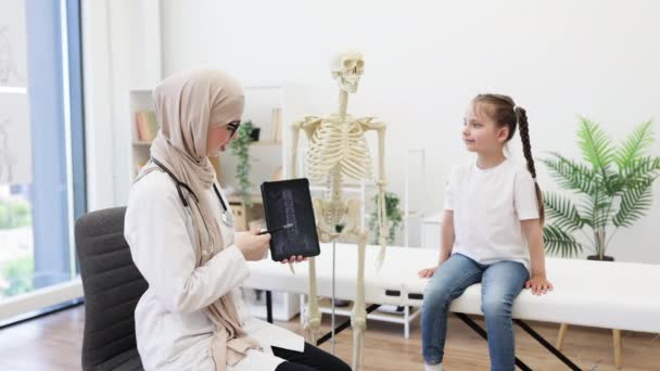 アラビアの小児科医は 医師の職場でタブレットと骨格にMri画像を使用して脊椎傷害について 慎重なガールに相談しています デジタルデータによる医療アドバイスを行うヒジャブの医療専門家 — ストック動画