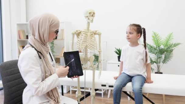 人間の骨格モデルに脊椎を検査する19人の慎重な子供が イスラム教徒の医師がデバイス上の画像検査を分析している ヒジャブの医療従事者が怪我治療に関する勧告を与える — ストック動画