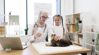 Doktorun ofisinde beyaz bir kızla kediyi okşayan tesettürlü, tesettürlü, pozitif Arap kadın. Yetişkin hayvanlara pire önleme protokolünü uyarlayan kedi uygulayıcısı.