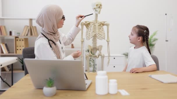 効率的なアラビアの専門家は 現代の診療所の医師のオフィスで好奇心旺盛な女子高生に骨格に肩のガードルを示しています 怪我の症状と治療を記述するヒジャブの関与した女性 — ストック動画