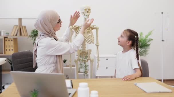 ヘッドスカーフのムスリム女性は 一般的な練習の医師のオフィスで解剖モデルの頭蓋骨の下顎を移動します 白いTシャツの白い女性は 仕事で骨格と楽しいセラピストを見ています — ストック動画
