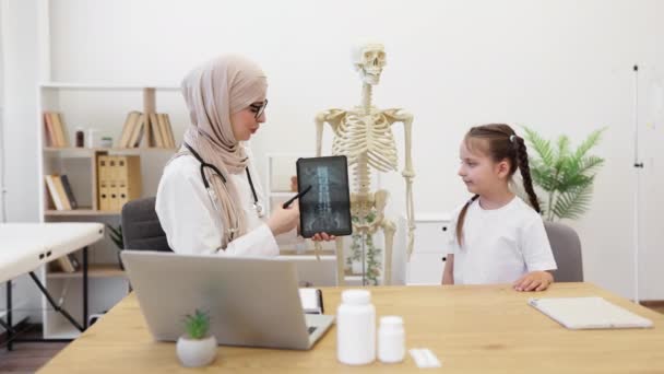 Ctスキャンでデジタルデバイスを保持するヒジャブのアラビアの医師と相互の視線を共有する慎重な女子高生の側面のビュー 脊髄損傷や病気に関する医療専門家による情報を得る — ストック動画