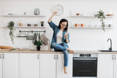Evde gülümseyerek cep telefonu ekranına bakarken sıcacık elbiseli çekici Asyalı kadın yumruğunu havaya kaldırıyor. Genç esmer bayan internette başarılı bir alışveriş yapmak için heyecanlı..