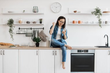 Gözlüklü neşeli Vietnamlı kadın bir ayak bileğiyle tezgahta oturuyor. Akıllı telefondan iş promosyonuyla e-posta alan duygusal genç bir kadın..