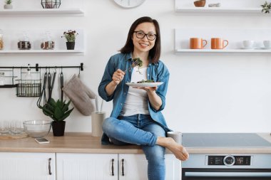 Çekici, çıplak ayaklı, rahat giyimli, salatalı bir kadın modern bir dairede tezgahta dinlenirken. Gözlüklü ince Asyalı kadın sağlıklı yaşam tarzını iyi beslenerek teşvik ediyor..
