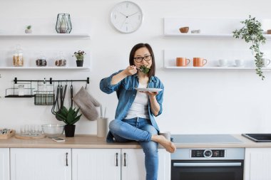 Çekici bir kadın şık bir mutfakta vejetaryen yemeğiyle tezgahta dinleniyor. Güzel görünümlü Asyalı kadın, boş zamanlarının sağlık yararlarıyla dolu salatayı kemiriyor..
