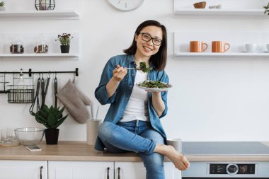 Çekici, çıplak ayaklı, rahat giyimli, salatalı bir kadın modern bir dairede tezgahta dinlenirken. Gözlüklü ince Asyalı kadın sağlıklı yaşam tarzını iyi beslenerek teşvik ediyor..