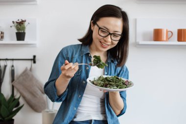 Japon kadının evde rahatlarken çatalla çatala bakıp gülümsemesini yakından izleyin. Kot gömlekli, vitamin ve besin yönünden zengin dengeli beslenme düzeniyle mutlu bir yetişkin..