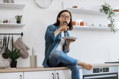 Dördüncü figürdeki Asyalı aşçı tezgahta oturup sabah taze salata yiyor. Sıkı çalışmaya başlamadan önce sağlıklı yiyeceklerin tadını çıkaran kot elbiseli, ince esmer bir yetişkin..