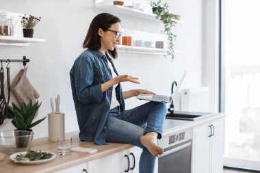 Asyalı bayanın çevrimiçi toplantıya katılırken dizleri üzerinde mutfak atölyesinde otururken görüntüsü. Girişim fikirlerini paylaşırken etkili iletişim becerileri gösteren şık serbest yazar.