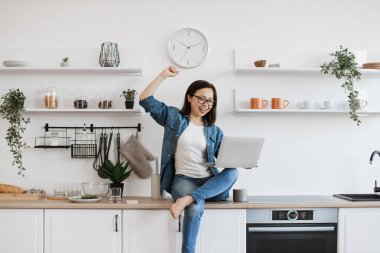 Mutfakta dizüstü bilgisayar ekranına bakarken zafer jesti yapan gözlüklü aşırı mutlu Çinli kadın. Kot elbiseli tutkulu iş kadını büyük bir galibiyetten sonra sevinç yaşıyor..