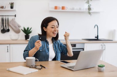 Ev içi bankamatik kartıyla dizüstü bilgisayar ekranına bakarken evet işareti yapan aşırı mutlu bir kadın. Mutlu Asyalı serbest yazar işvereninden maaş aldıktan sonra heyecanlanıyor.