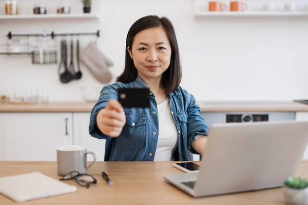 stock image Focus on smiling asian lady reaching out credit card while sitting at kitchen table with gadgets on surface. Cheerful freelancer promoting cashless transactions via home banking service for customers.