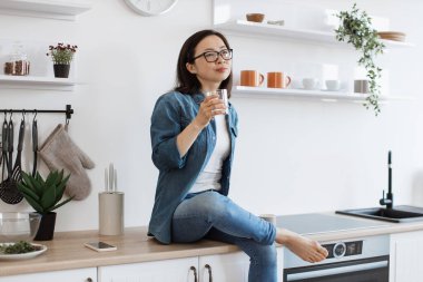Evde ahşap tezgahta dinlenirken içi sıvı dolu bir bardakla hayal kuran çıplak ayaklı kadın. Gözlüklü kaygısız Asyalı yaz akşamı için planlar yapıyor..