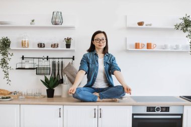 Modern mutfağın tezgahında nilüfer çiçeği pozu veren günlük kıyafetler ve bardaklar içindeki neşeli Asyalı kadın. Evde zihinsel ve duygusal stresi azaltmak için yoga yapan mutlu zayıf kadın..