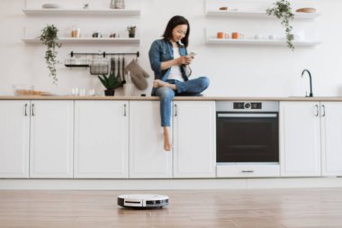 Genç Asyalı kadın mutfak tezgahında bacak bacak bacak üstüne atmış otururken kişisel cep telefonundan robot elektrik süpürgesini izliyor. Evde modern aletlerle kolay temizlik..