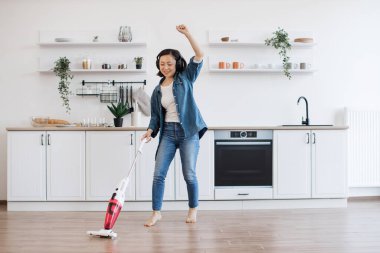 Gülümseyen Vietnamlı kadın kablosuz süpürgeyle yerleri temizlerken kulaklıkla neşeli bir müzik eşliğinde kıpırdıyor. Evde bakım yaparken elektrikli aletlerle eğlenen güzel bir ev hanımı..