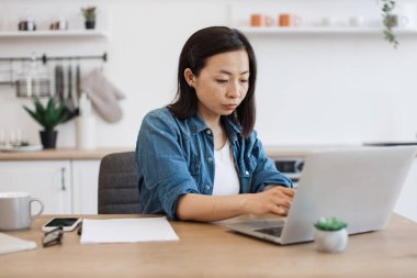 Mutfakta kalırken, rahat kıyafetli, taşınabilir dijital cihaz kullanan çekici bir kadının yan görüntüsü. Düşünceli Japon kadın araştırma projesi için online kütüphaneden kitap indiriyor..