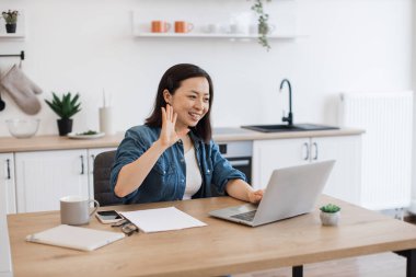 Çekici Asyalı kadın kişisel bilgisayarında online konferans sırasında el sallıyor. Koyu renk kısa saçlı genç bayan rahat dairesinde video görüşmesi yapıyor. Tasarımcı mutfağının arka planı.