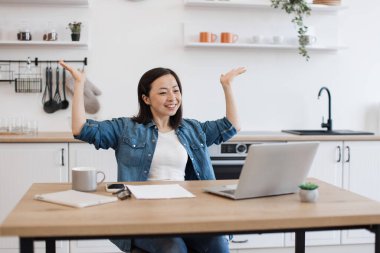 Aşırı neşeli Asyalı öğretim görevlisi, mutfaktaki ahşap masada otururken, akıllı teknolojiler vasıtasıyla çevrimiçi webinar sunuyor. Heyecanlı genç öğretmen yeni iş planıyla ilgili canlı yayın yapıyor..