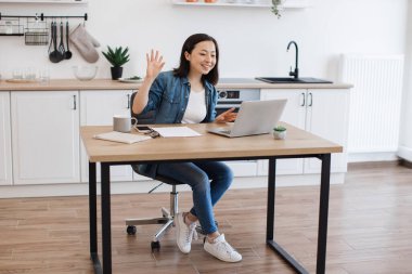 Asyalı bir kadının iş arkadaşıyla internet konferansı sırasında ahşap masada otururken konuştuğu geniş açı. Ev işyerindeki iş geliştirme hakkında beyin fırtınası yapan çekici serbest yazar.