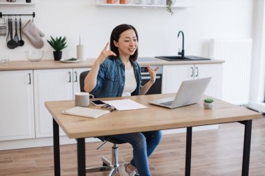 Asyalı bir kadının iş arkadaşıyla internet konferansı sırasında ahşap masada otururken konuştuğu geniş açı. Ev işyerindeki iş geliştirme hakkında beyin fırtınası yapan çekici serbest yazar.