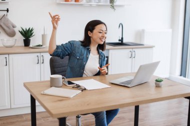 Asyalı bir kadının iş arkadaşıyla internet konferansı sırasında ahşap masada otururken konuştuğu geniş açı. Ev işyerindeki iş geliştirme hakkında beyin fırtınası yapan çekici serbest yazar.