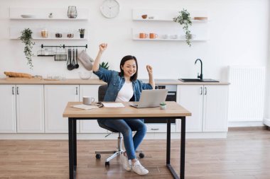 Neşeli Asyalı kadın modern laptopta iyi haberler okurken olumlu duygularını ifade ediyor. Evde mutlu mesut dolaşan, gündelik kıyafetli, pozitif genç bir kadın..