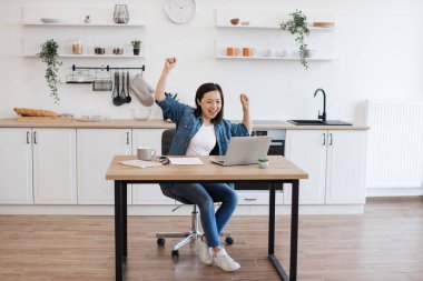 Neşeli Asyalı kadın modern laptopta iyi haberler okurken olumlu duygularını ifade ediyor. Evde mutlu mesut dolaşan, gündelik kıyafetli, pozitif genç bir kadın..