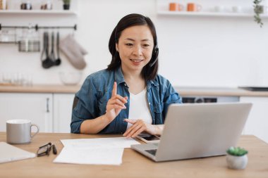 Günlük kıyafetlerle giyinmiş çekici Asyalı kadın kişisel bilgisayardaki video konuşmalarıyla bir şeyler anlatıyor. Evdeki online toplantı sırasında parmağıyla işaret ederek gülümseyen genç yönetici..