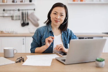 Kulaklıklı Asyalı kadın ve kot gömlekli kadının modern dairede dizüstü bilgisayarın arkasında mesai saatlerini geçirdiği yakın plan. Uzman danışman evden iş konularında uzman tavsiyesi veriyor.