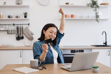 Mutfak masasında bilgisayarla kredi kartını tutarken zafer jesti yapan mutlu Asyalı kadın. Başarılı alışverişi kutlayan heyecanlı kadın bankacılık uygulaması ile online ödeme.