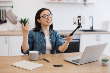 Enerjik kadın kişisel blogu için modern akıllı telefonuyla selfie çubuğuyla kendini filme çekiyor. Konforlu oturma odasında kablosuz dizüstü bilgisayar kullanırken gözlüklü Asyalı bir kadının online kurslar kaydetmesi.