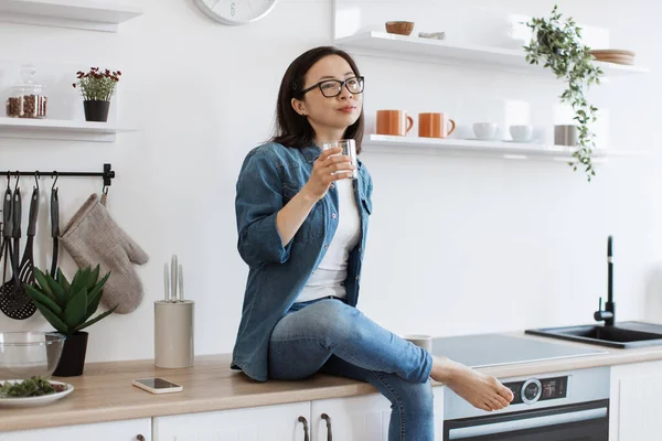 stock image Relaxed barefoot lady in jeans daydreaming with glass full of liquid while resting on wooden countertop at home. Carefree asian person in eyewear making plans for summer evening in kitchen interior.