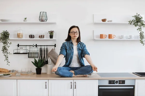 Peaceful asian woman meditating while sitting on white designer kitchen set. Slender female dressed in casual clothes practicing yoga to achive life balance and harmony at cozy apartment.