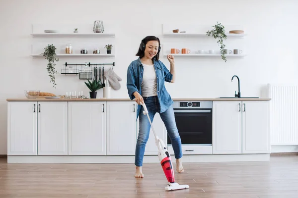 stock image Joyful young using cordless vacuum cleaner and lmodern headphones during housework. Active asian woman in denim wear enjoying favorite music and dancing at kitchen interior.