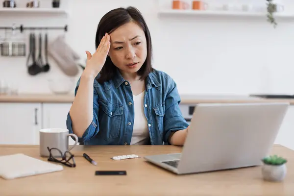 Feverish asian lady in denim clothes feeling forehead while looking at laptop camera in kitchen of bright apartment. Unwell person with medicine arranging virtual appointment with health professional.