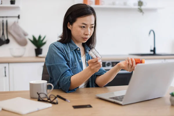Young asian lady with medicine in hands getting online consultation from general practitioner via laptop. Unhealthy freelancer asking advice about treatment from stuffy nose during allergy season.