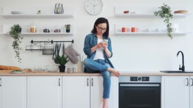 Genç Asyalı kadın ahşap mutfak mobilyalarının üzerinde oturuyor ellerinde cep telefonu ve düşünceli bir şekilde yana bakıyor. Sevimli genç bir kadın rahat bir dairede otururken arkadaşlarıyla sohbet ediyor..