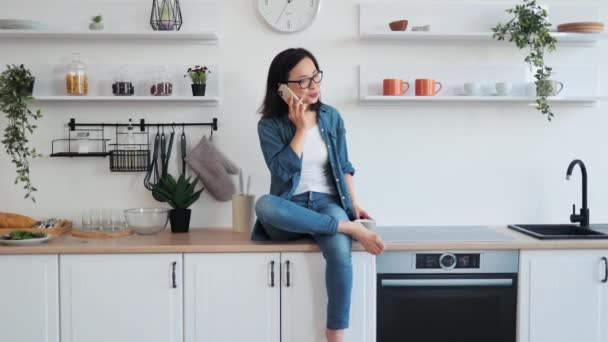 デザイナーのキッチンで休憩する魅力的なアジアの女性は 手でホットティーのカップで現代のスマートフォンで話しながらセットします 自宅でガジェットを使用して楽しい会話を楽しむ眼鏡のスリム女性 — ストック動画