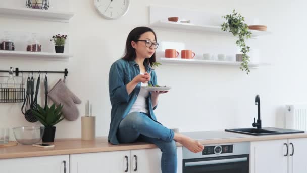 キッチンインテリアの別の膝の上に1足首と座っている間 若いアジア料理ベジタリアン料理 晴れた週末にゼロから自家製の食事を作成するメガネの健康的なスリム女性 — ストック動画
