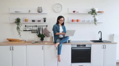 Asyalı bir kadının dizinde bilgisayarla şık bir mutfakta atölyesinde rahat bir şekilde oturuşu. Gözlüklü genç bir öğrenci evdeki yeni kariyer seçenekleriyle ilgili online makale okuyor..